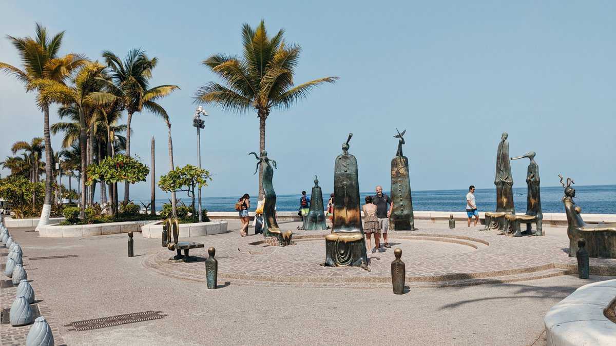 Large square with palm trees and a lot of bronze statues facing the beach