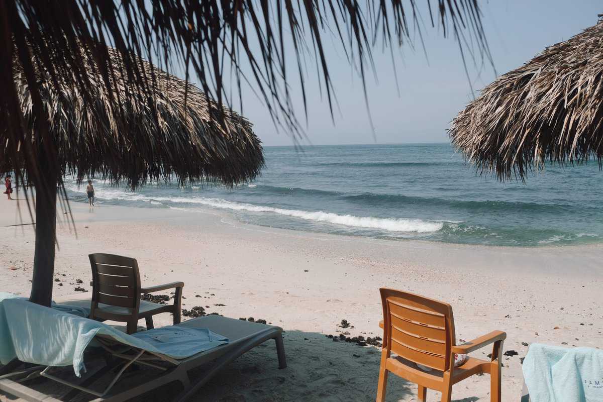 Beach chairs and a thatched umbrella on a sandy beach with clear blue water in the background.